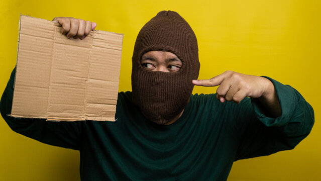 Young Asian male wearing brown balaclava holds blank corrugated board while pointing index finger towards it against bright yellow backdrop. The masked individual gazes sideways at
