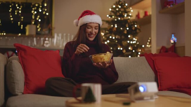Woman in Santa hat eating potato chips while enjoying Christmas movie, 4K