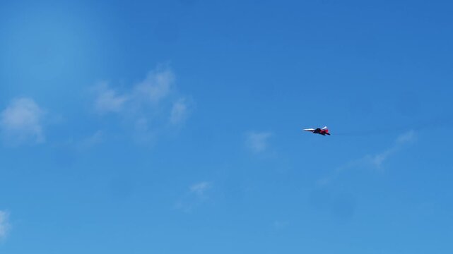 Single military fighter jet painted in bright red white and blue tricolor scheme flies across clear blue sky. Supersonic combat aircraft leaves faint smoke trail