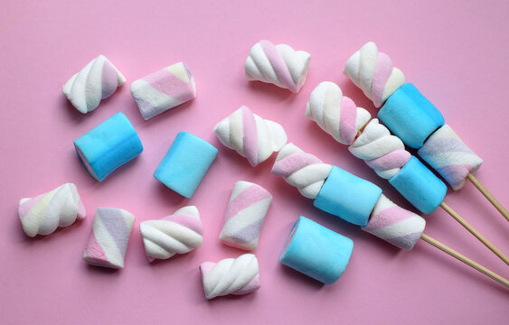 Multicolored marshmallows are skewered on wooden sticks against a pink background.