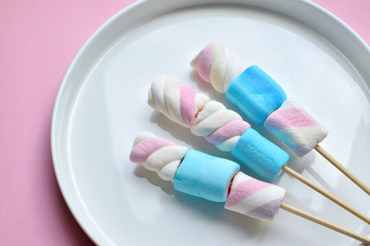 Multicolored marshmallows skewered on wooden sticks on a white plate. Copy space.