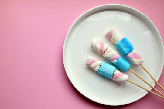 Multicolored marshmallows skewered on wooden sticks on a white plate. Copy space.