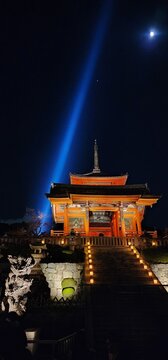 Dramatic vertical night view looking up stone stairs toward the illuminated vermilion entrance gate (Kyozo or Saimon) at Kiyomizu-dera, with the iconic blue beam of light shooting into the dark night.