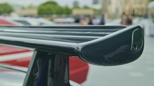 Detailed shallow depth of field shot focusing on a glossy carbon fiber rear wing spoiler on a tuned sports car, with blurred people walking in the background.