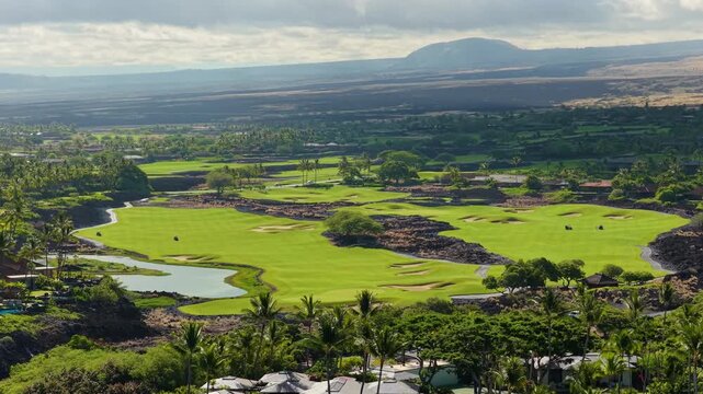 Drone aerial view of vibrant green golf course fairways on Big Island Hawaii surrounded by black lava fields palm trees and luxury resort villas with volcanic mountain on the horizon.