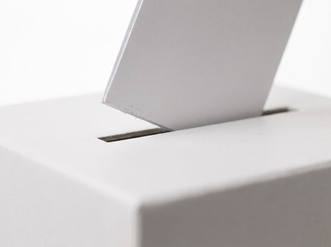 A close-up shot of a white ballot box with a white ballot being inserted into the slot, symbolizing voting and democratic process.