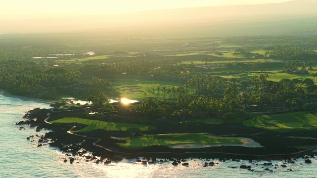 Drone aerial of luxury oceanfront golf course on Big Island Hawaii at golden sunset with palm trees lava rock coastline turquoise ocean waves green fairways and resort villas along shore.