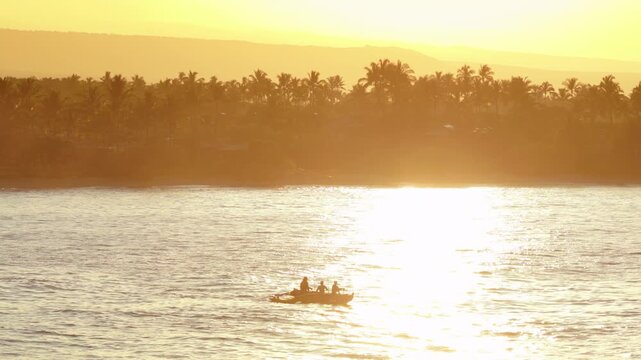 Silhouetted outrigger canoe with paddlers gliding across shimmering golden ocean water at sunset off Big Island Hawaii with palm trees volcanic slopes and warm tropical light on coastline.