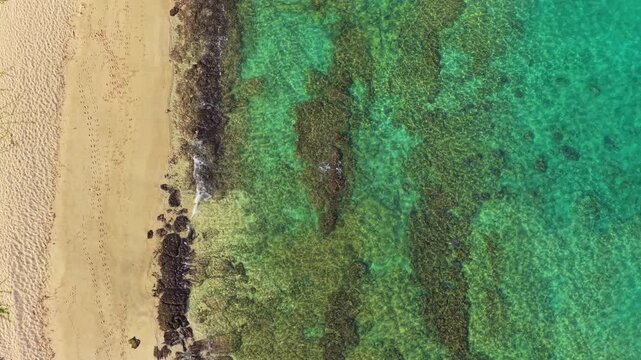 Aerial drone view over crystal clear turquoise water and coral reef along a golden sandy beach on the Big Island of Hawaii with footprints in sand and gentle waves washing over lava rocks.