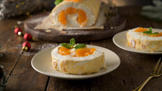 A slice of delicate meringue roulade filled with cream and bright tangerine segments rests on a ceramic plate, garnished with fresh mint leaves under warm holiday lighting.