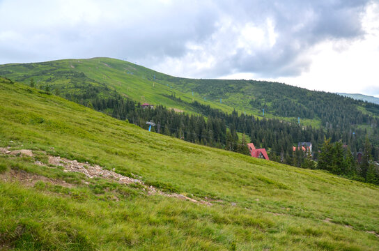Vast green mountain slopes feature scattered evergreen trees, ski lift towers, and small red-roofed buildings under a cloudy overcast sky. Dragobrat, Carpathians, Ukraine