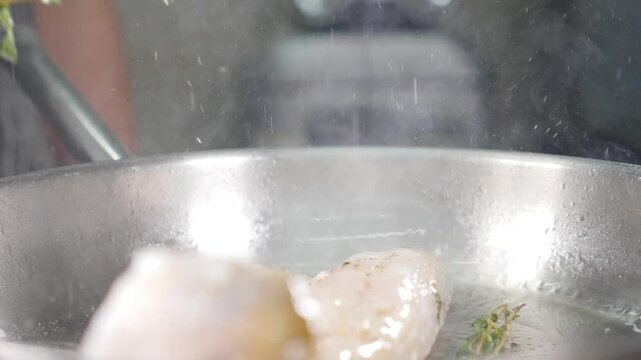 Close-up of white fish fillet cooking in stainless steel pan with fresh thyme sprig. Rising steam, hot oil, professional kitchen seafood preparation, healthy gourmet meal Slow motion 4k video