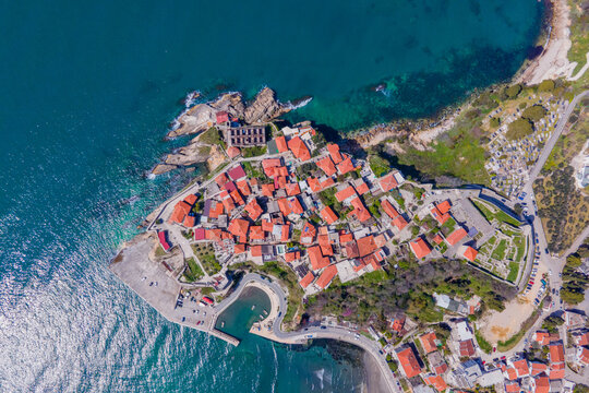 A drone view shows a small coastal town on a rocky peninsula with red tiled roofs surrounded by turquoise sea, roads and a marina, a scenic travel destination.
