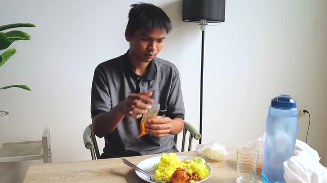 Young Asian man adds chili sauce to turmeric rice while eating crackers at the dining table