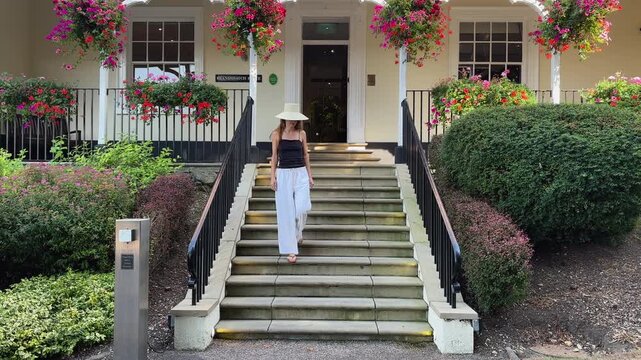 A young woman in a sunhat and summer outfit walks down the front steps of a grand hotel, surrounded by lush gardens and vibrant hanging flowers.