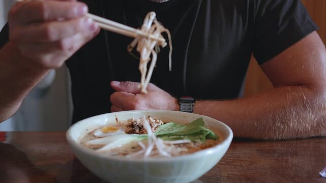 Man with short hair enjoys a bowl of noodles with chopsticks, featuring broth, vegetables, and meat, while seated at a wooden table in a casual dining environment
