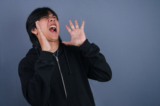 Young Asian Man Screaming In Despair With Open Mouth And Hands Up In Studio Setting