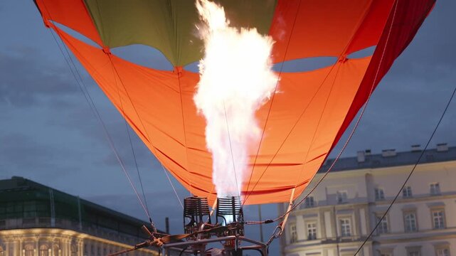 Colorful hot air balloon fills with air as flames ignite from the burner, creating a striking visual against the evening sky in a city