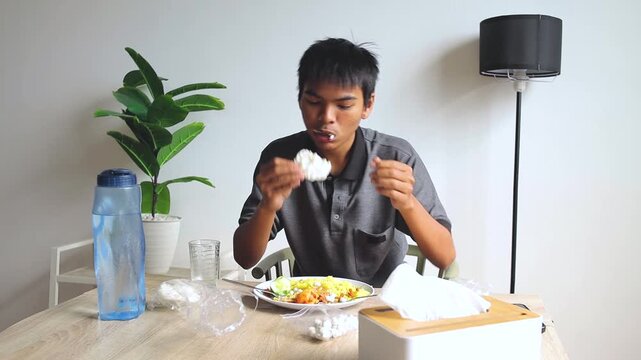 Young Asian man taking bite crispy crackers while eating turmeric rice at dining table