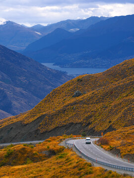 A campervan driving down a winding hairpin bend on Crown Range Road with layers of autumn mountains and Lake backdrop, South Island, New Zealand