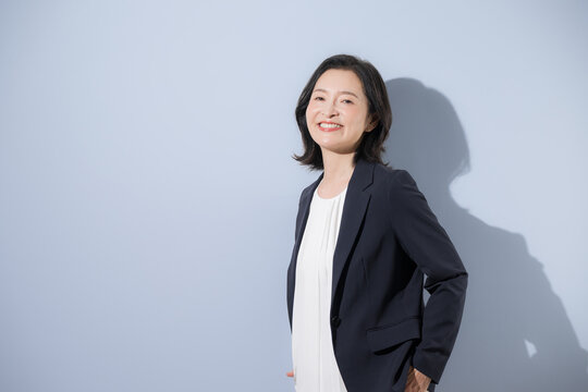 A middle-aged Japanese woman standing with a confident smile; a female businessperson in her 50s wearing a suit, with her hands in her pockets, looking directly at the camera