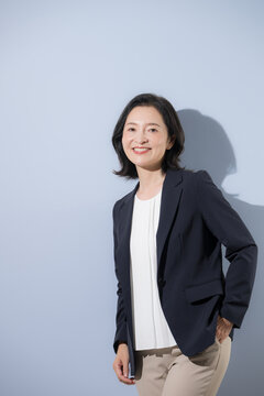 [水色背景]自信のある笑顔で立つ日本人ミドル女性、スーツ姿でポケットに手を入れる50代の女性ビジネスパーソンカメラ目線