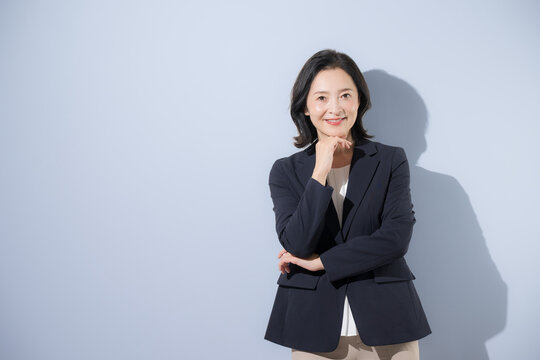 [水色背景]仕事について考える日本人ミドル女性、腕を組み顎に手を添えて考える50代の女性ビジネスパーソンカメラ目線上半身