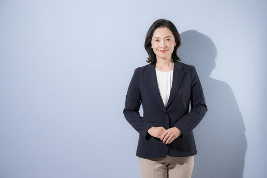 [Light blue background] A middle-aged Japanese woman in a suit, standing with her hands clasped and facing the camera&mdash;a portrait of a trustworthy female business professional in her 50s