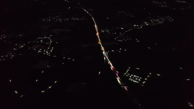Cinematic aerial: Night drone tracking a long line of vehicle light trails curving through the dark countryside, passing by a small rural settlement.