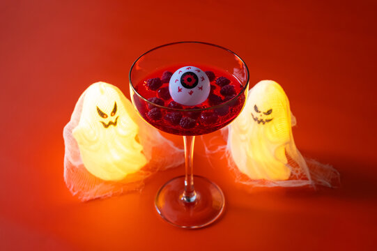 Halloween cocktail with an eyeball garnish and berries, flanked by glowing ghosts on an orange background.