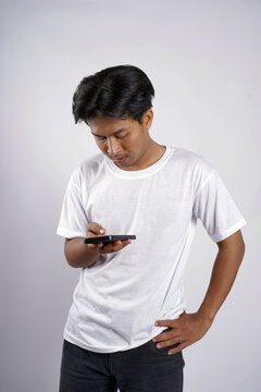 Young man wears white t-shirt, stares at phone. He holds device, leans slightly, hand on hip. Focused, perhaps searching or confused by content. Perfect for tech, lifestyle, or social media themes