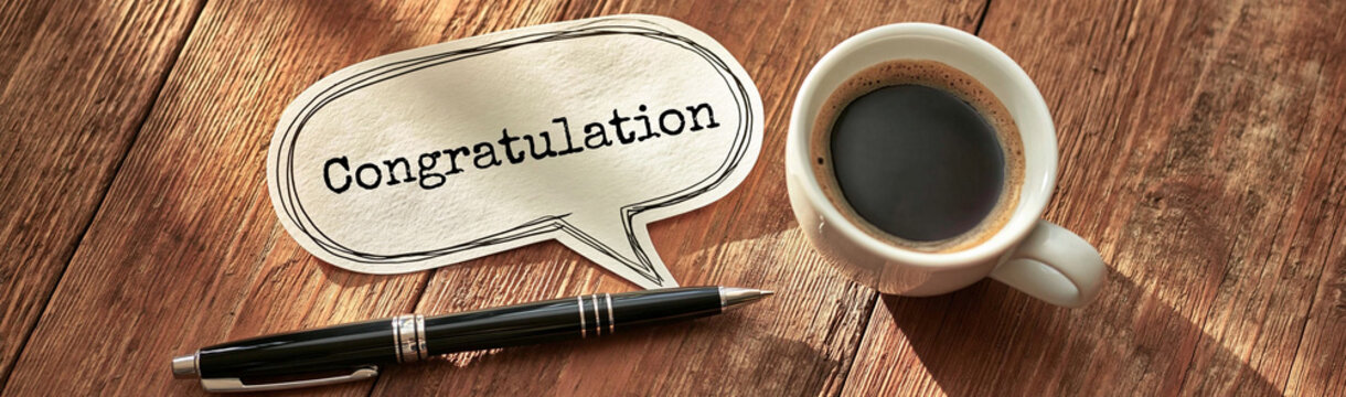 A Moment of Celebration: A cup of hot coffee sits beside a pen and a speech bubble conveying Congratulation, set on a rustic wooden surface, ideal for conveying well wishes and acknowledging success.