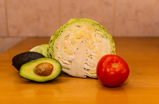 Variedad de vegetales crudos: medio repollo, un tomate rojo y una palta cortada a la mitad mostrando su carozo, dispuestos sobre una mesa de madera.