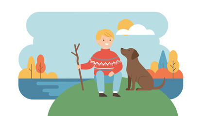 Fototapeta premium Young boy holding a wooden stick sits on a grassy hill with his loyal brown dog in a scenic autumn landscape.