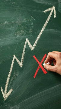 Graph Downturn Reversed: A hand draws a red cross on a chalkboard, marking the end of a downward trend, as an arrow curves upward, signifying a resurgence or turnaround.