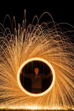 Man spins burning steel wool in dark night. Long exposure light painting creates bright orange fire circle. Glowing sparks fly in spiral motion. Person performs pyrotechnic art show with glowing