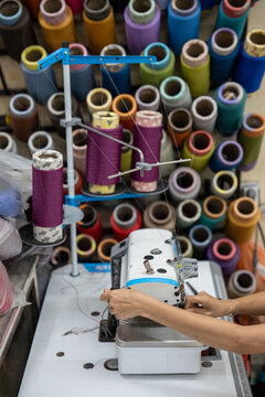 Weaving production in Vietnam, female seamstress hands operating sewing machine in workshop, professional woman sewer fixating thread on machine at worktable surrounded by colorful thread spools.