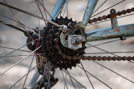 rusty steel bicycle frame close-up, highlights a multi-speed gear cassette, severely rusted drive chain, metallic spokes, and weathered axle parts showing aging patina on a blurred background