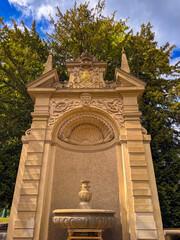Hercules Fountain in Prague's Royal Garden, built in 1670. The Baroque sandstone structure features a decorative niche, a stone basin, an imperial coat of arms, and green trees behind. © pijav4uk