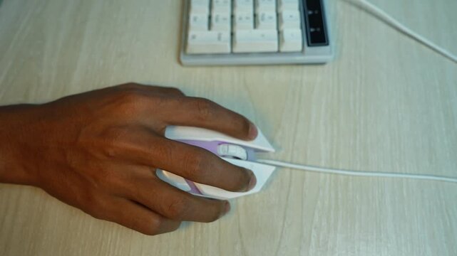 Hand using computer mouse beside keyboard, Close-up of a person's hand using a white computer mouse beside a keyboard on a light wooden desk.