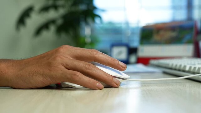 Hand using computer mouse at office desk, Close-up of a man's hand using a computer mouse on a desk in a bright office, with keyboard, monitor and plant softly blurred in the background.