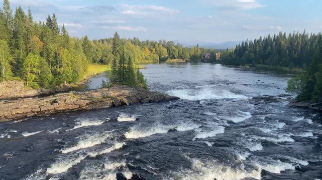 A wide river with a strong current, surrounded by trees and rocks under a blue sky, J&auml;mtland, Sweden