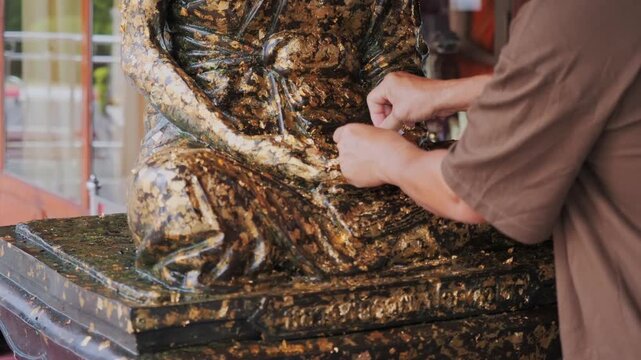 Close up hands applying gold leaf on buddha statue base during buddhist merit making ritual