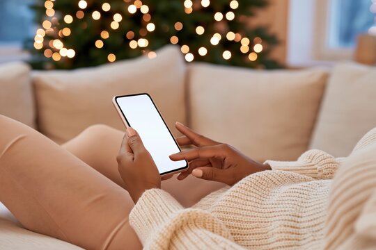 Woman in cozy knit sweater using smartphone on beige sofa with blurred christmas tree lights in background