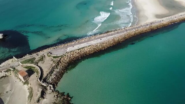 Aerial view of a stone breakwater extending into turquoise ocean waters beside a sandy beach and coastal town with mountains in the distance in Tarifa, Spain