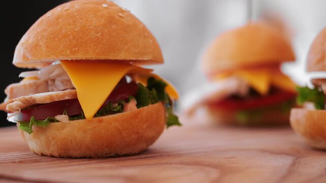 Close-up of mini burger with sliced meat, cheese, tomato and lettuce in a soft bun, served on a wooden board with other burgers blurred in background.