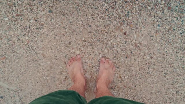 Male feet standing on clear water and sandy beach, top-down view. 4K
