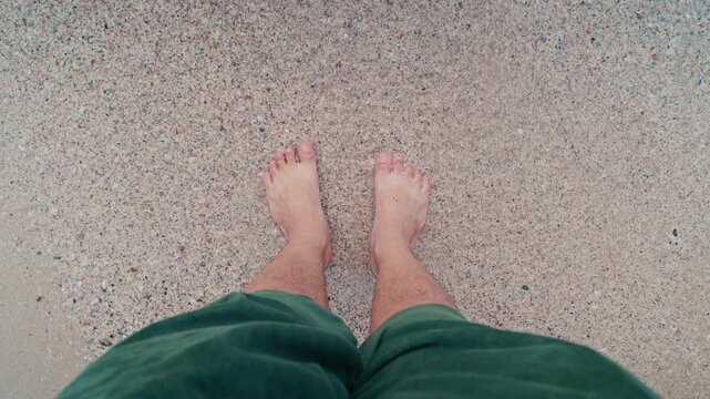 Male feet standing on clear water and sandy beach, top-down view. 4K