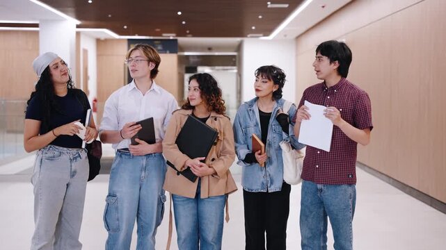 Wide shot of five diverse university students standing in a campus hallway while a classmate speaks holding a sheet of paper