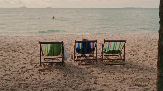 4K Rear view of a person relaxing on a colorful beach chair looking out at the calm ocean with a paddleboarder in the distance